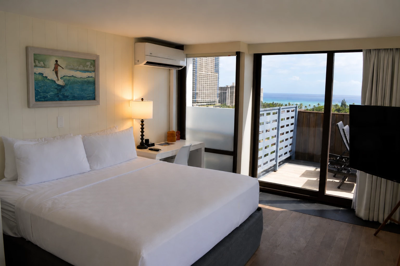 A hotel room with a white bed, desk, lamp, and artwork, featuring a balcony with a city and ocean view.