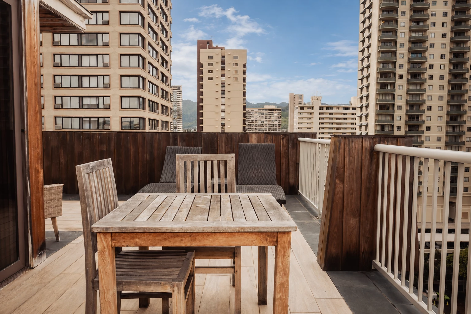 A balcony with a wooden table and chairs, overlooking a cityscape with mountains in the distance.
