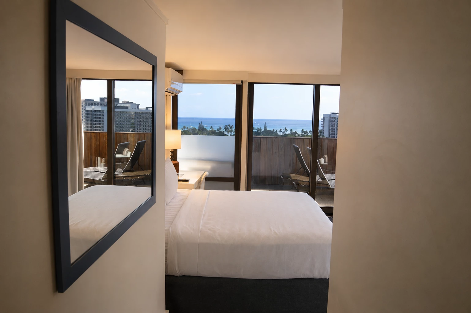 A hotel room with a bed, mirror, and balcony overlooking the ocean.