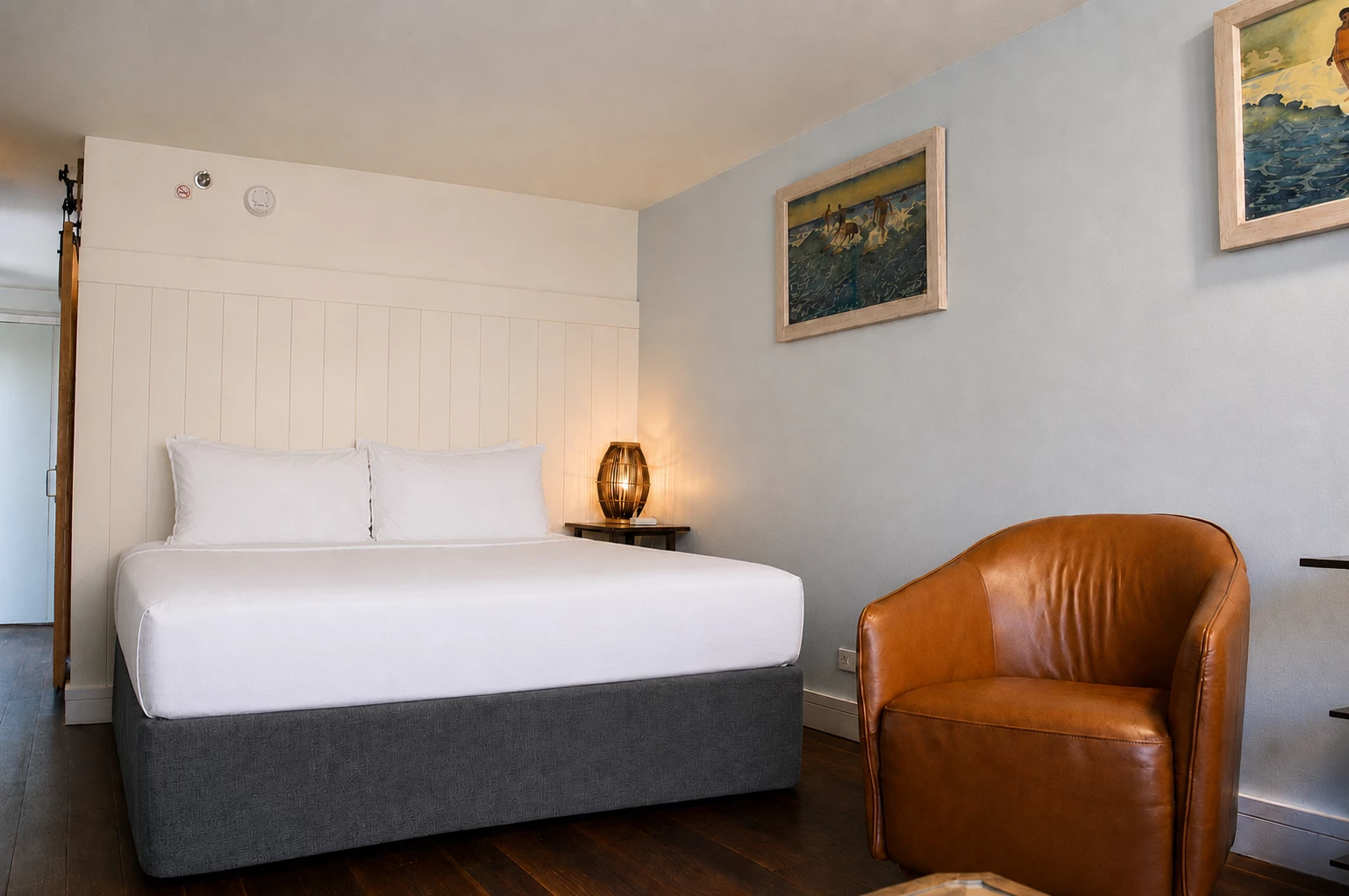 A hotel room with a bed, chair, lamp, and artwork on the walls.