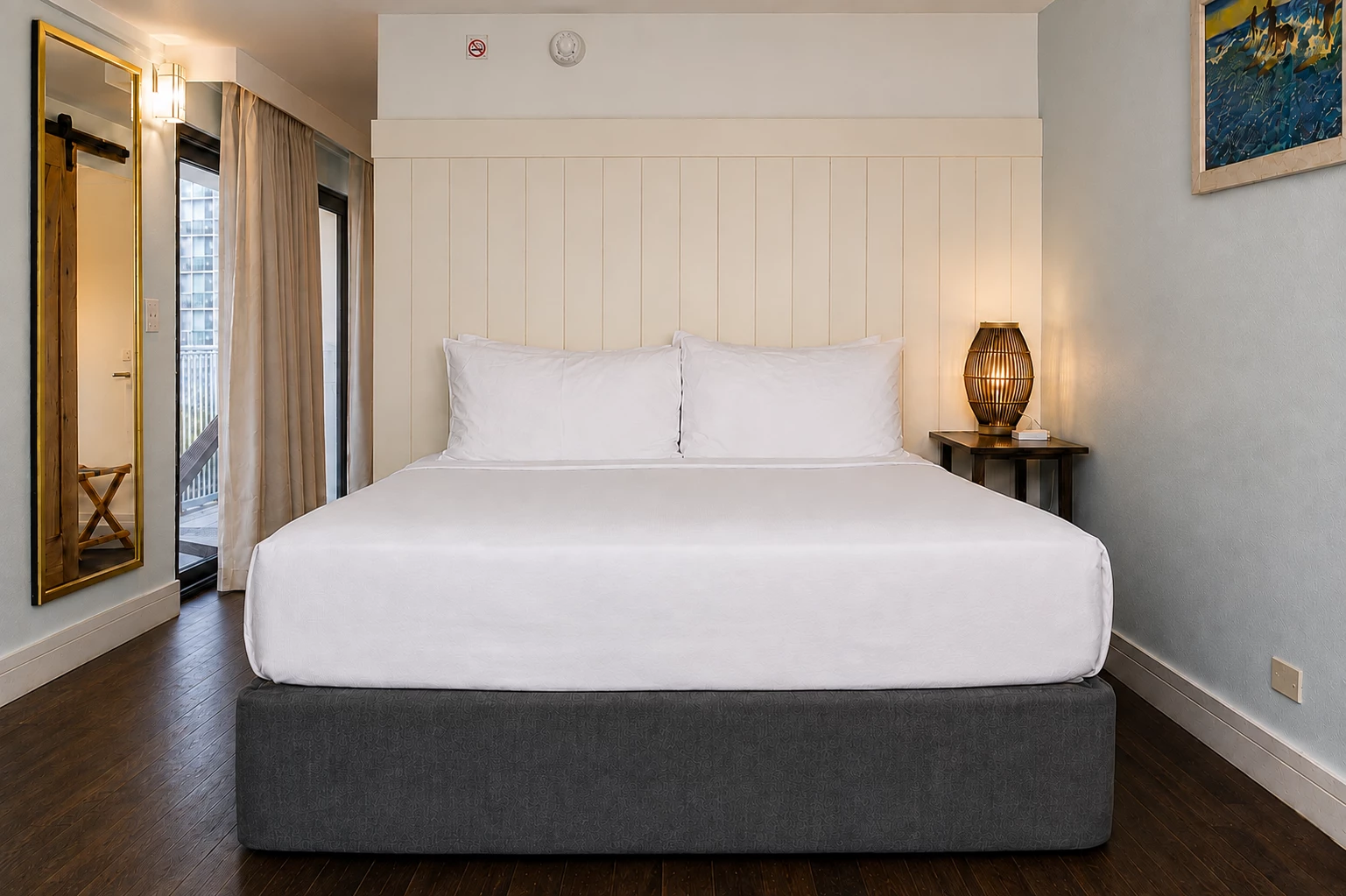 A neatly made bed with white linens and pillows, a nightstand with a lamp, and a mirror on the wall in a hotel room.