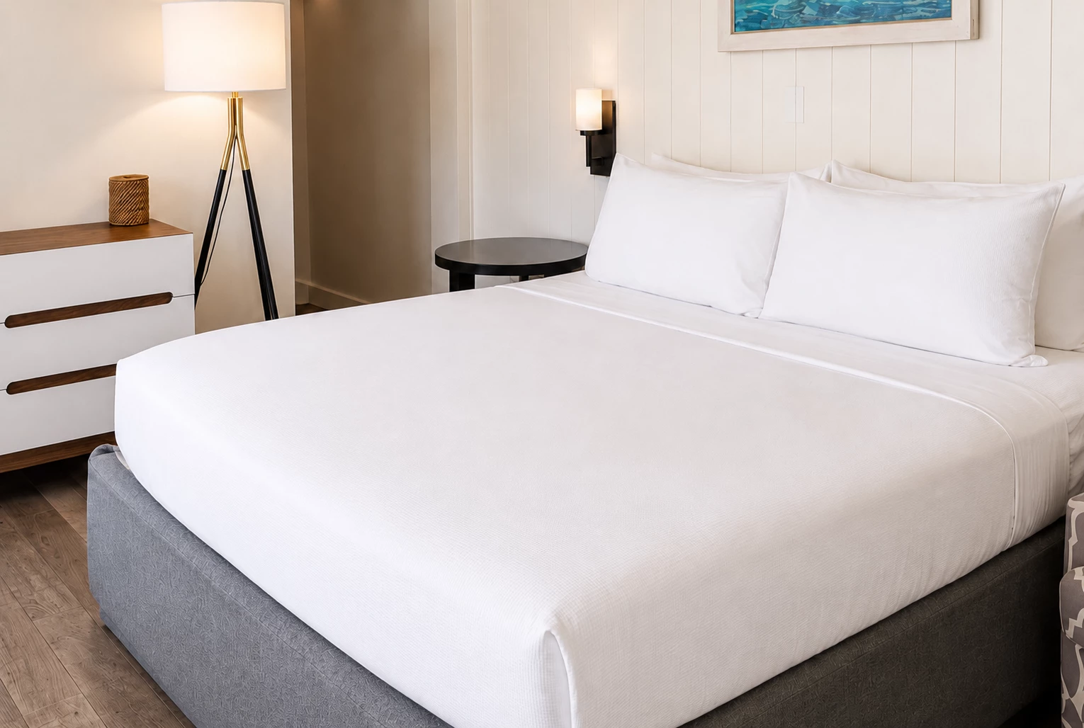 A neatly made bed with white linens and pillows in a room with a nightstand and lamp.