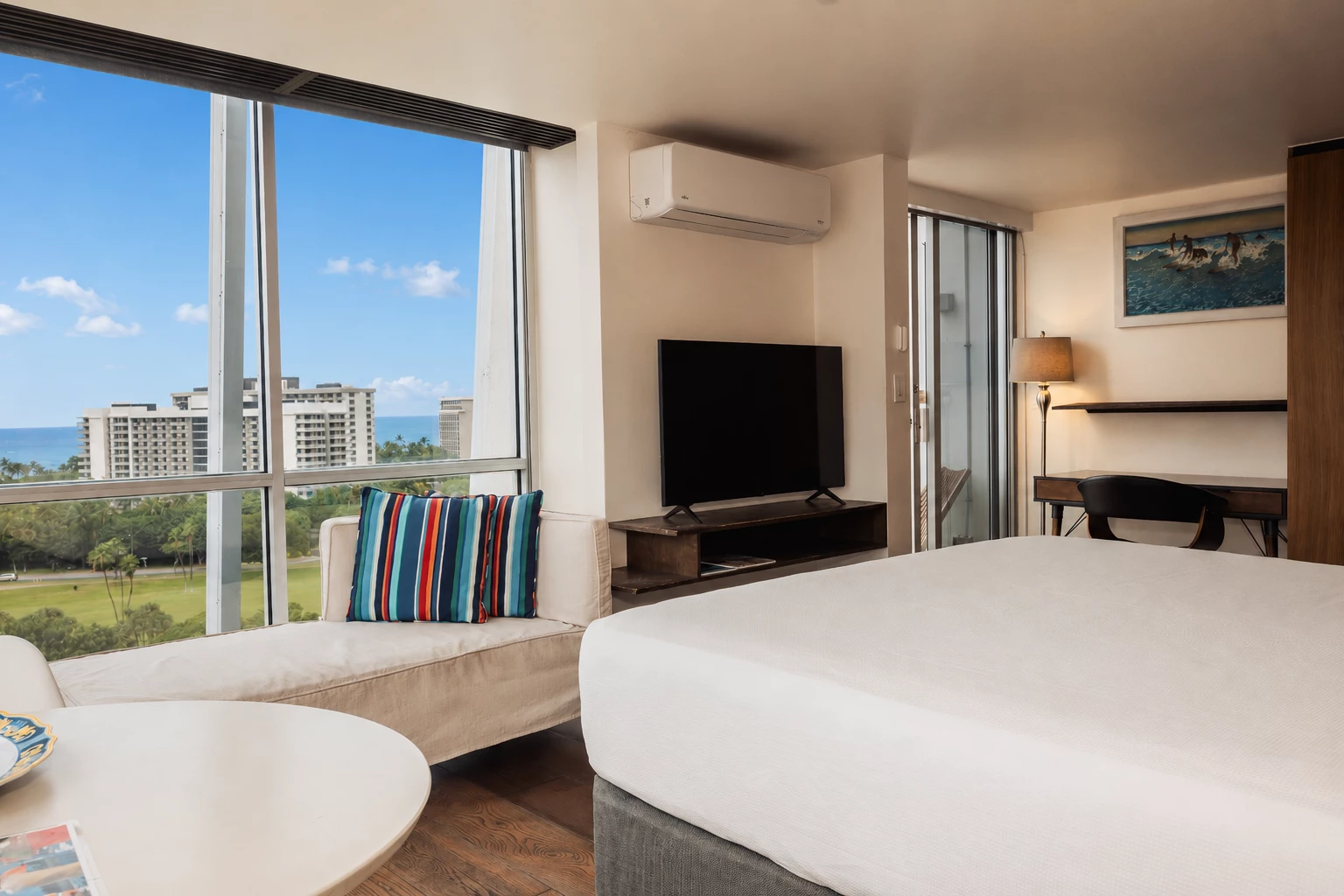 A hotel room with a large window, a bed, a sofa, a TV, and a desk.
