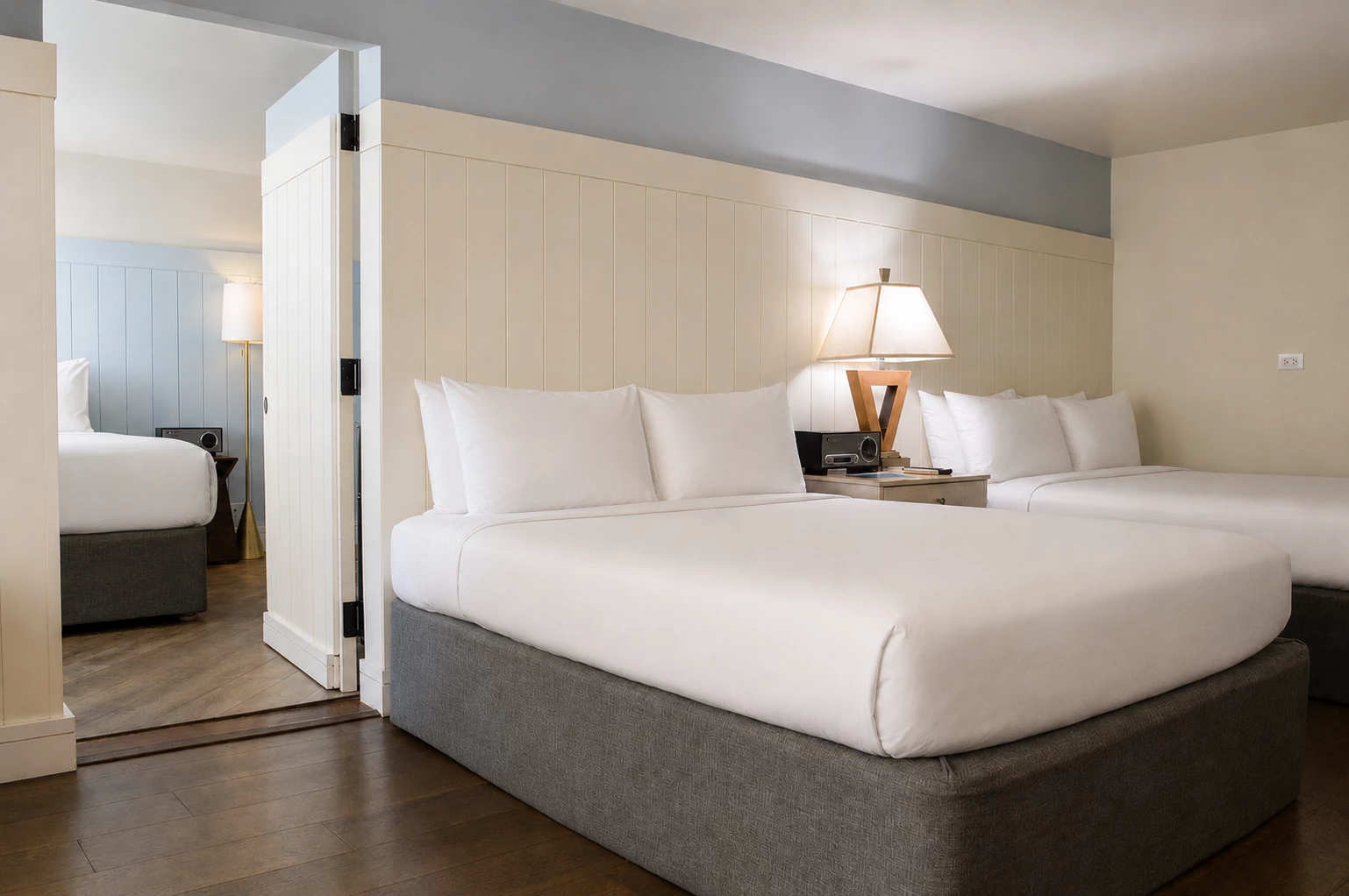 A hotel room with two double beds, a wood floor, and a divider wall with a mirror.