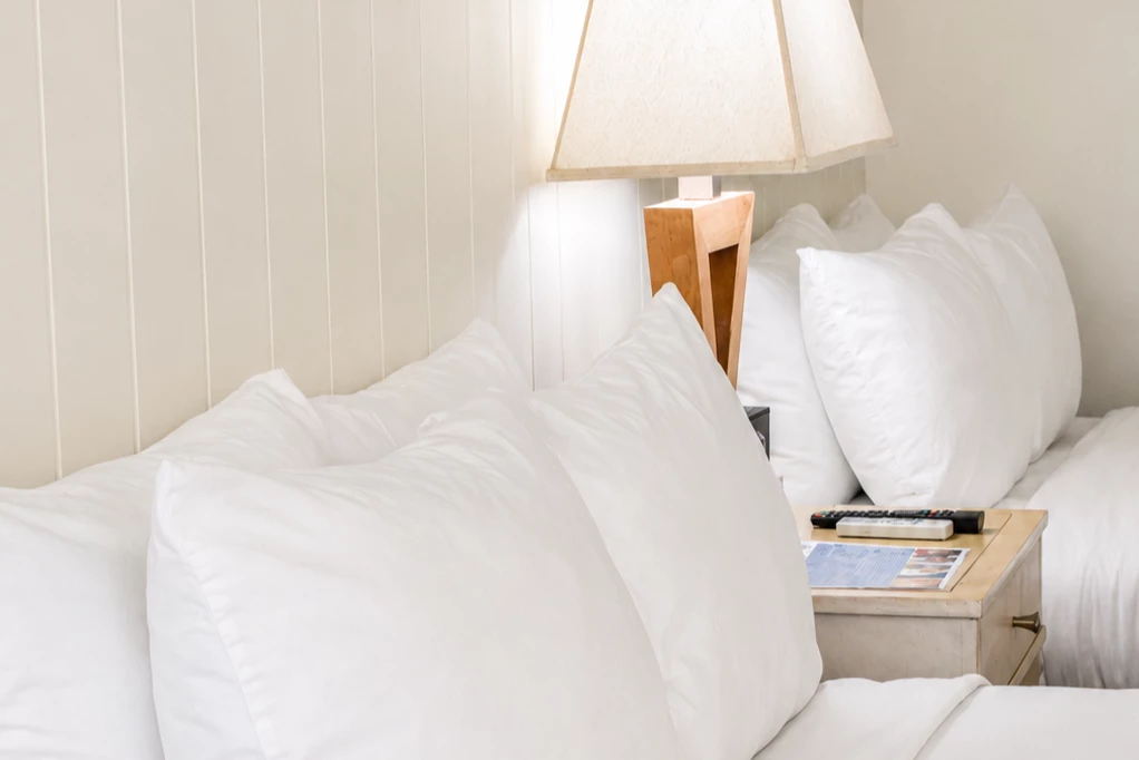 A close-up view of a bed with white bedding and pillows, a wooden nightstand with a lamp and remote control.
