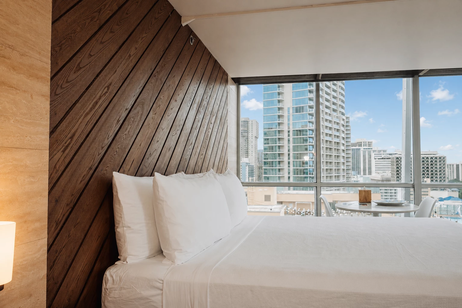 A hotel room with a bed, table, and chairs in front of a window showing a city view.