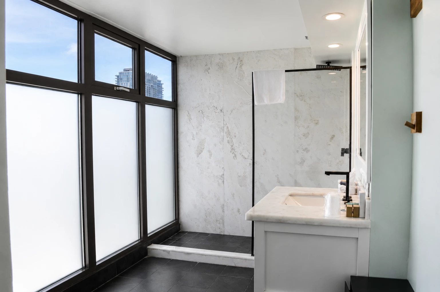 A modern bathroom with a glass-enclosed shower, marble countertop, and a window with a city view.