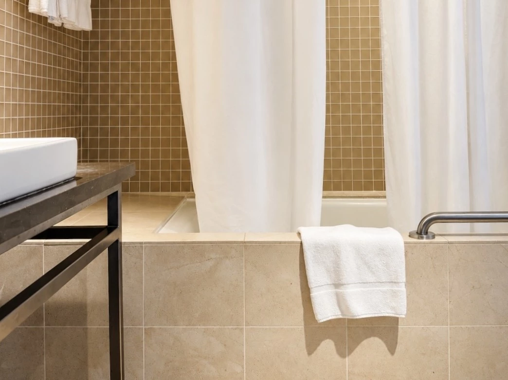a bathroom with a tan tile shower and bathtub with a white curtain, a white sink, and a white towel hanging on the side of the tub