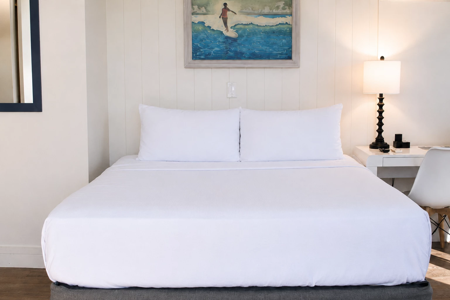 A neatly made bed with white linens and two pillows in a room with a desk, lamp, and artwork.