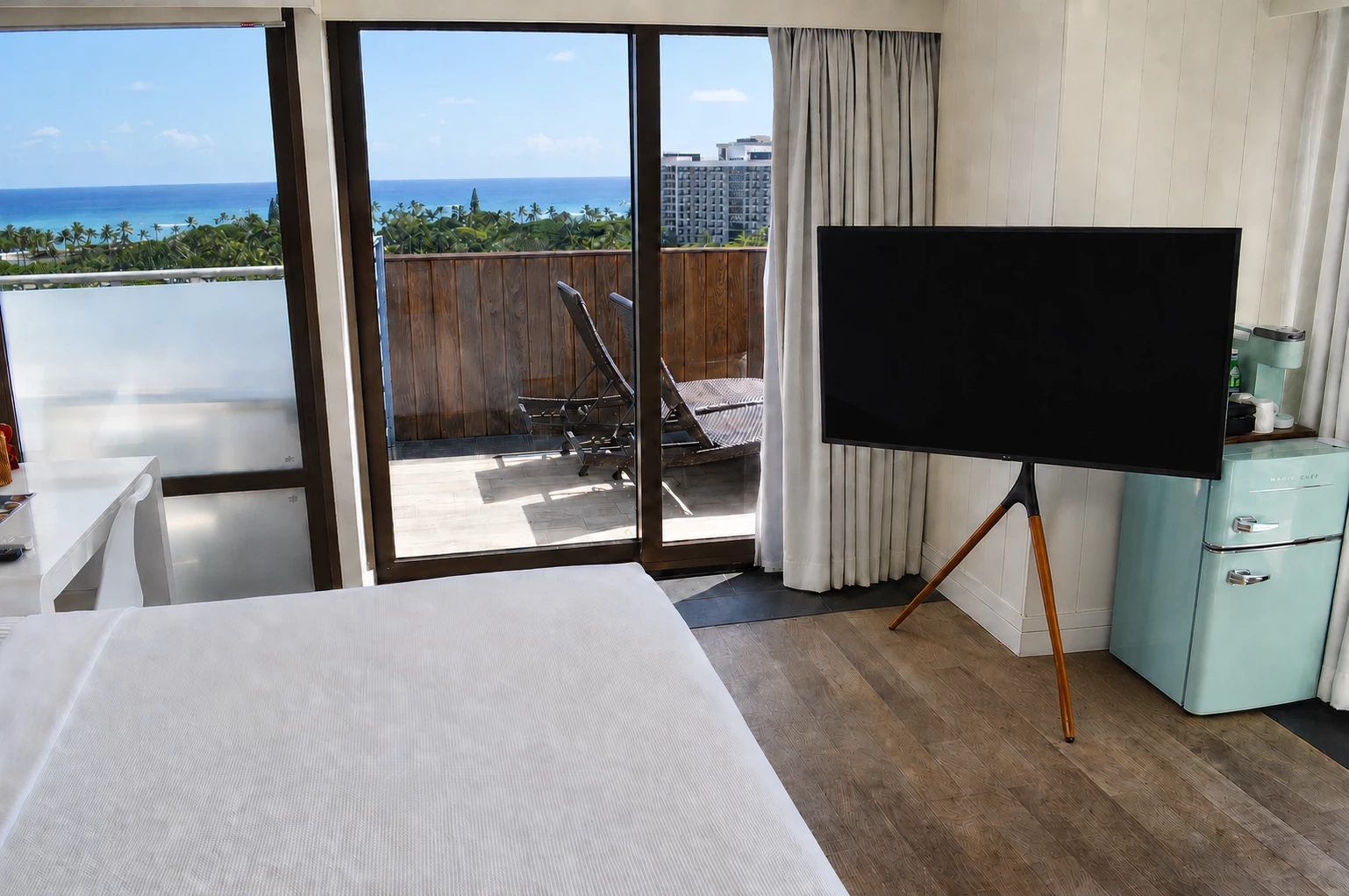 A hotel room with a bed, TV, mini-fridge, and balcony with an ocean view.