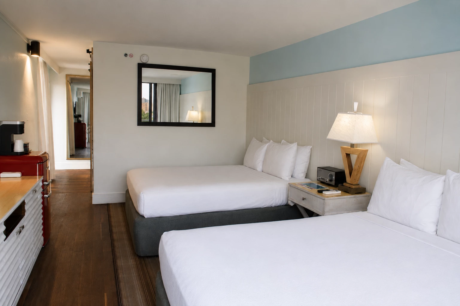 A hotel room with two beds, a nightstand, a lamp, a mirror, and a refrigerator.