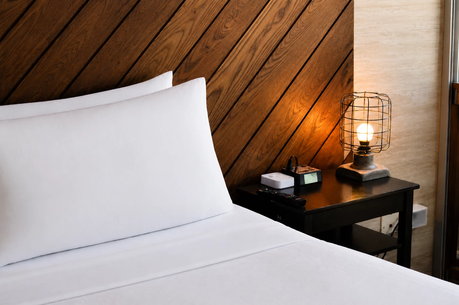 A close-up of a bed with a wooden headboard, white bedding, and a nightstand with a lamp.
