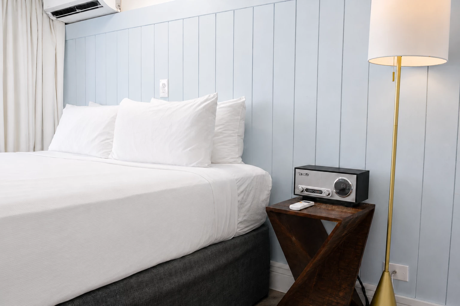 A neatly made bed with white linens and pillows against a light blue paneled wall, with a wooden nightstand, a radio, and a floor lamp.