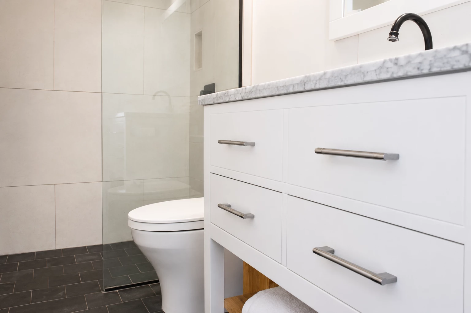 A modern bathroom with a white vanity, marble countertop, glass-enclosed shower, and dark tile floor.
