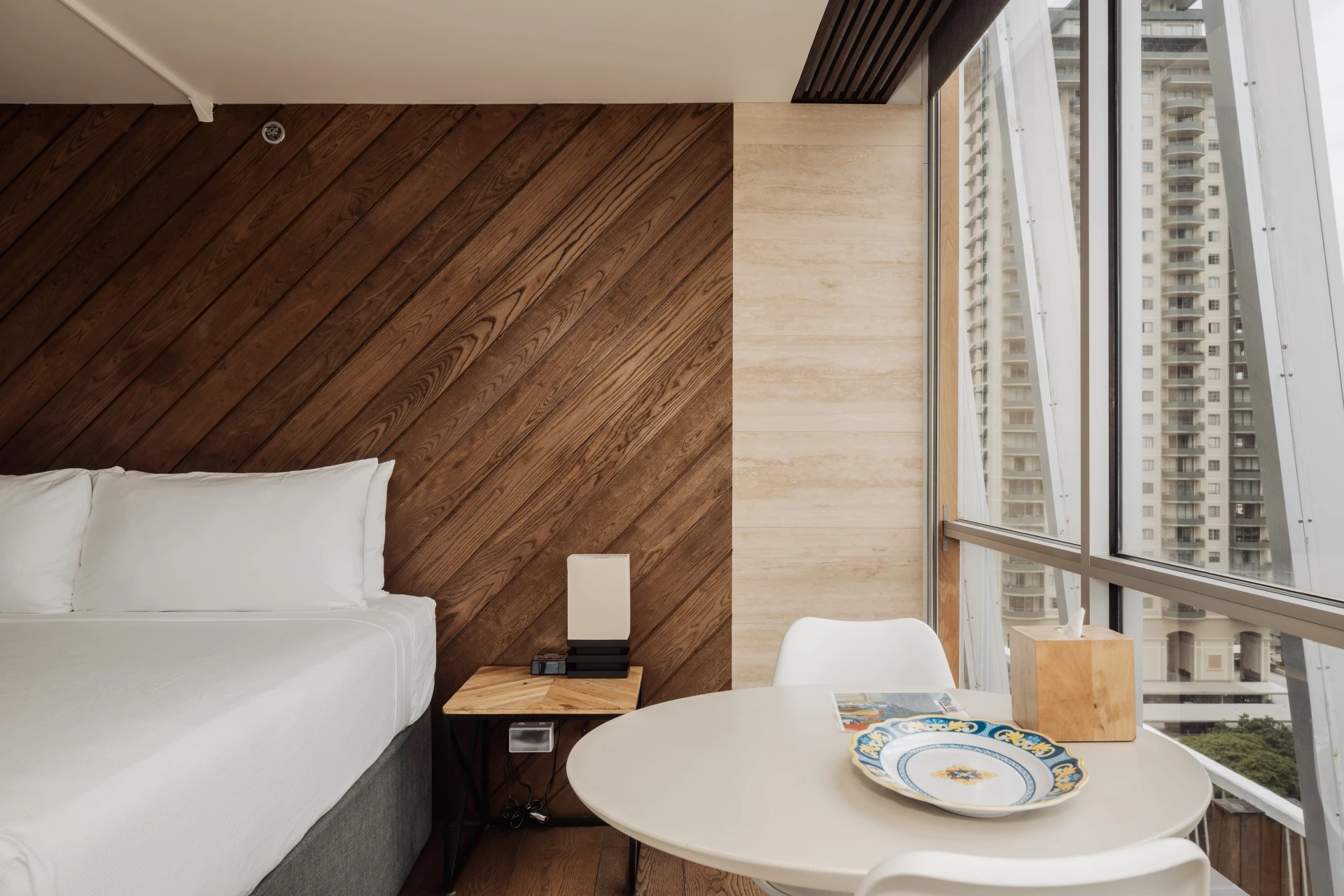 A hotel room with a bed, table, chair, and window showing a city view.