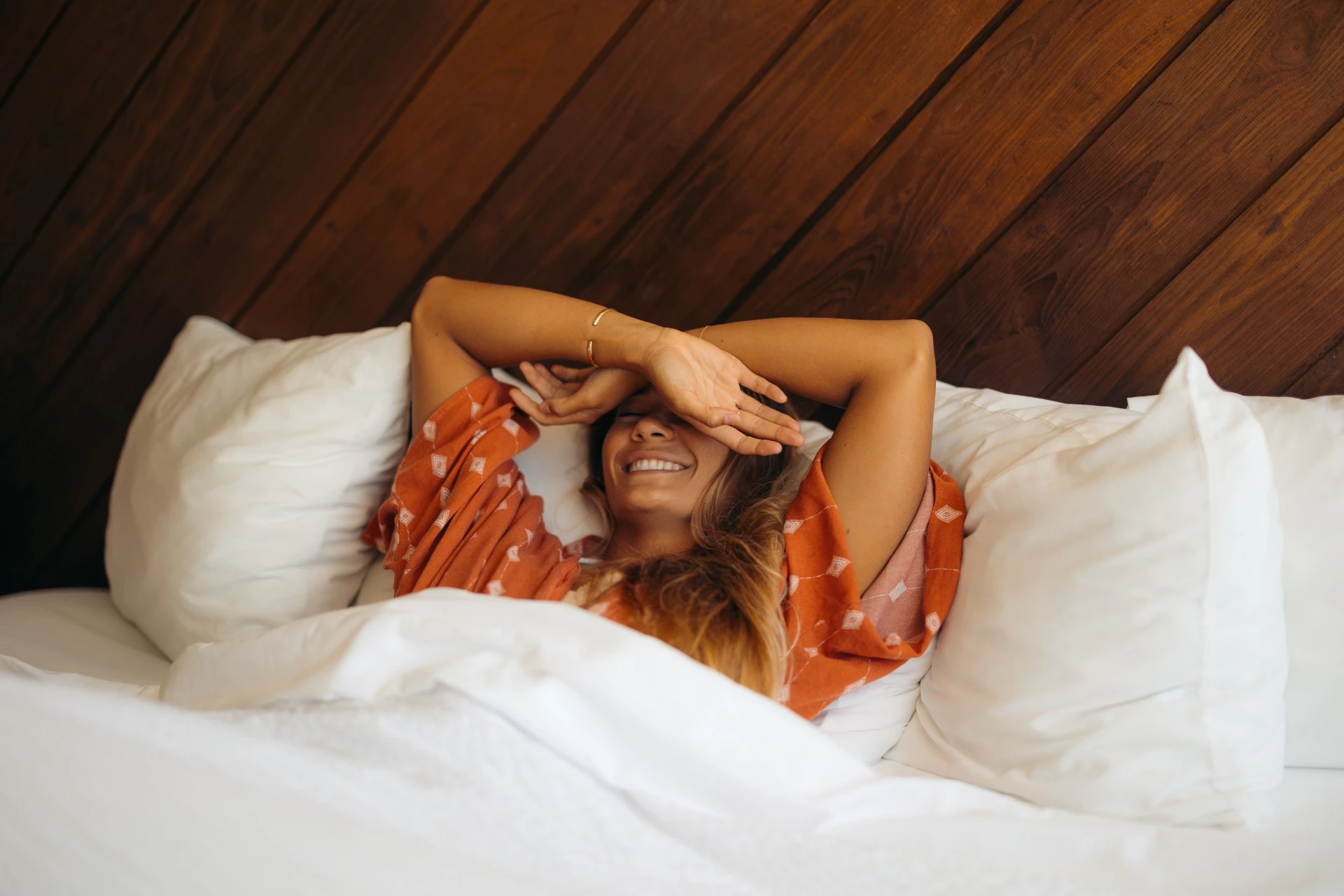 A woman in an orange dress lies on her back in bed with her hands above her head.