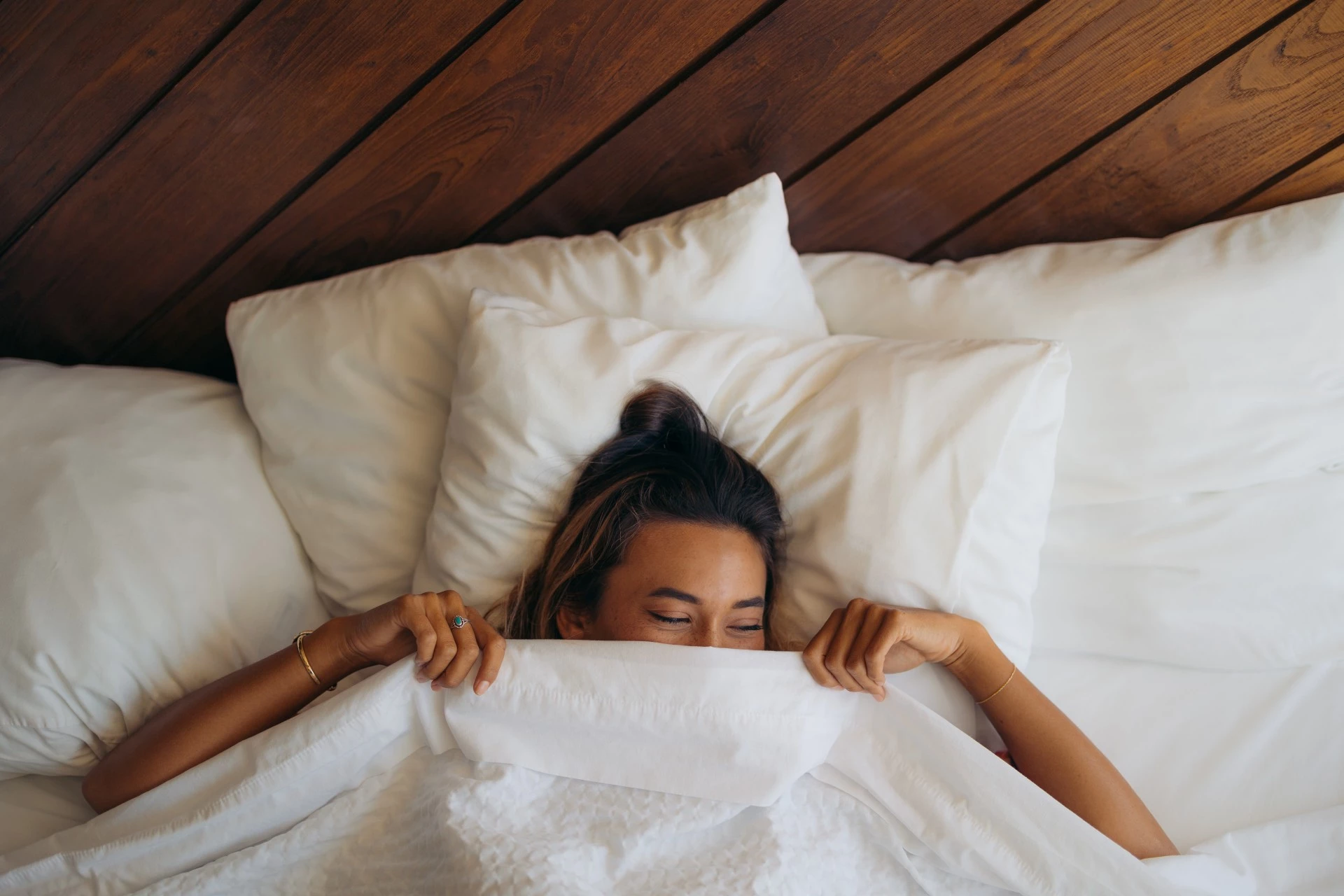 A woman lies in bed with white pillows and bedding, and a wood-paneled ceiling above her.
