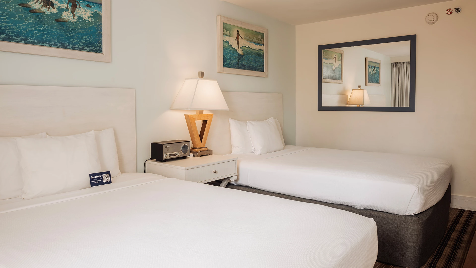A hotel room with two beds, a nightstand, lamp, mirror, and artwork on the walls.