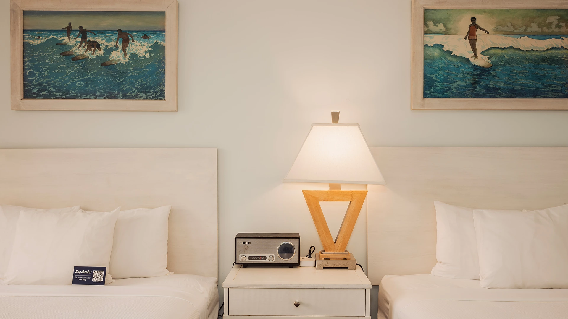 A hotel room with two beds, a nightstand with a lamp and radio, and surf-themed artwork on the wall.
