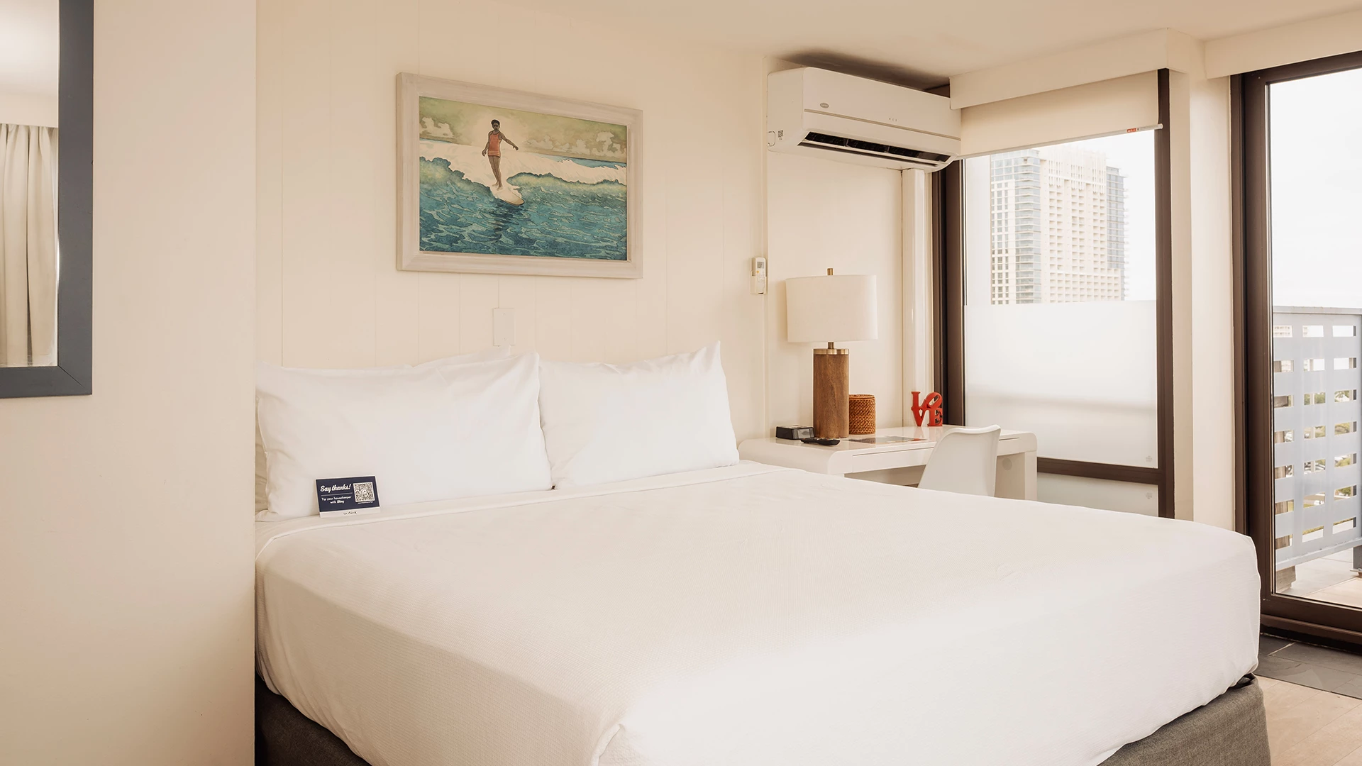 A hotel room with a large bed, a window with a balcony, a desk with a lamp, and a painting on the wall.