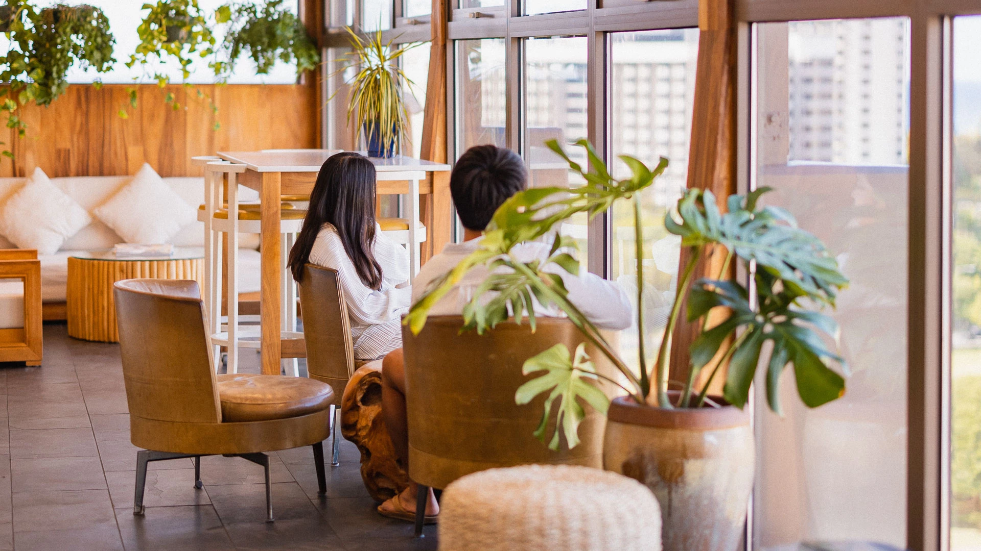 A lounge area with a sofa, chairs, tables, and plants in front of a large window.