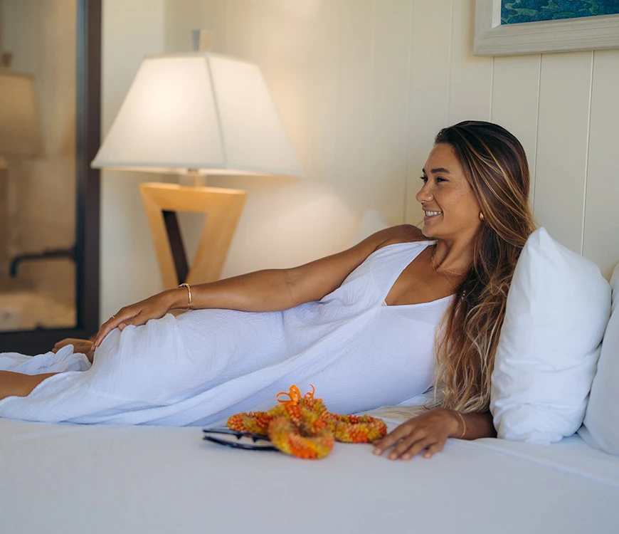 A woman in a white dress lying on a bed with a lamp and pillows.