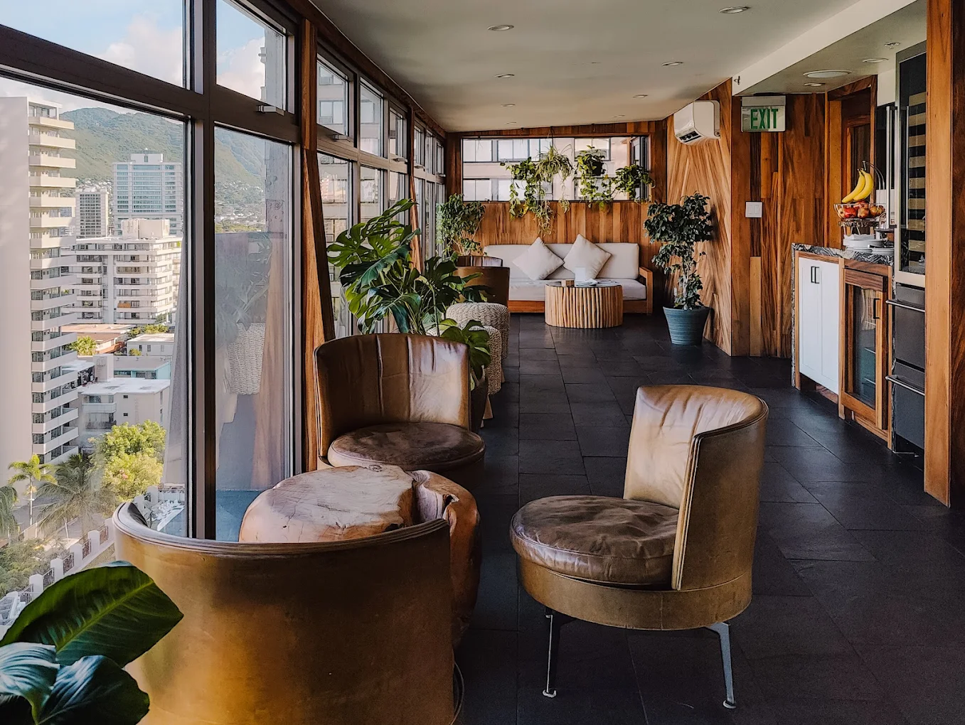 A stylish lounge area with leather chairs, wood paneling, and plants, offering a city view.