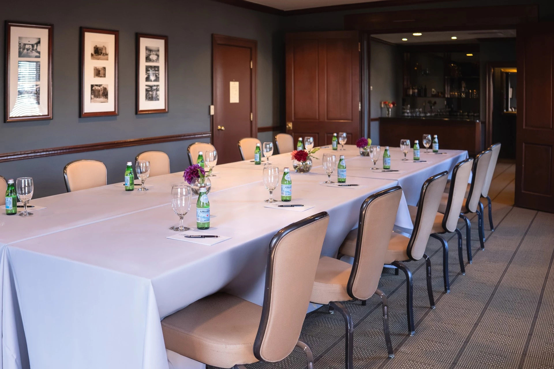 A long table set for a meeting or event in a room with gray walls and a bar in the background.