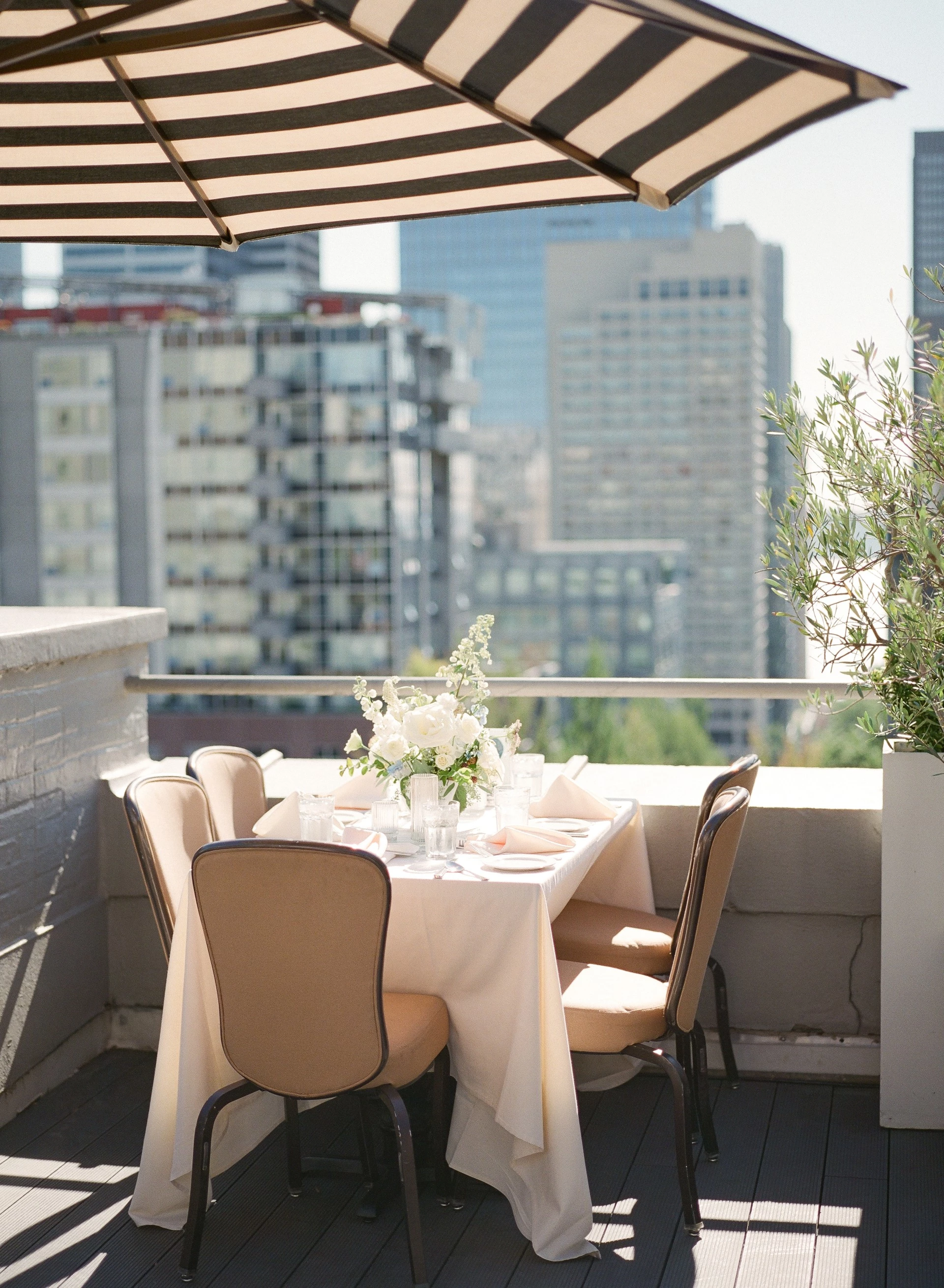 A dining table with chairs and a umbrella on a rooftop patio overlooking the city.
