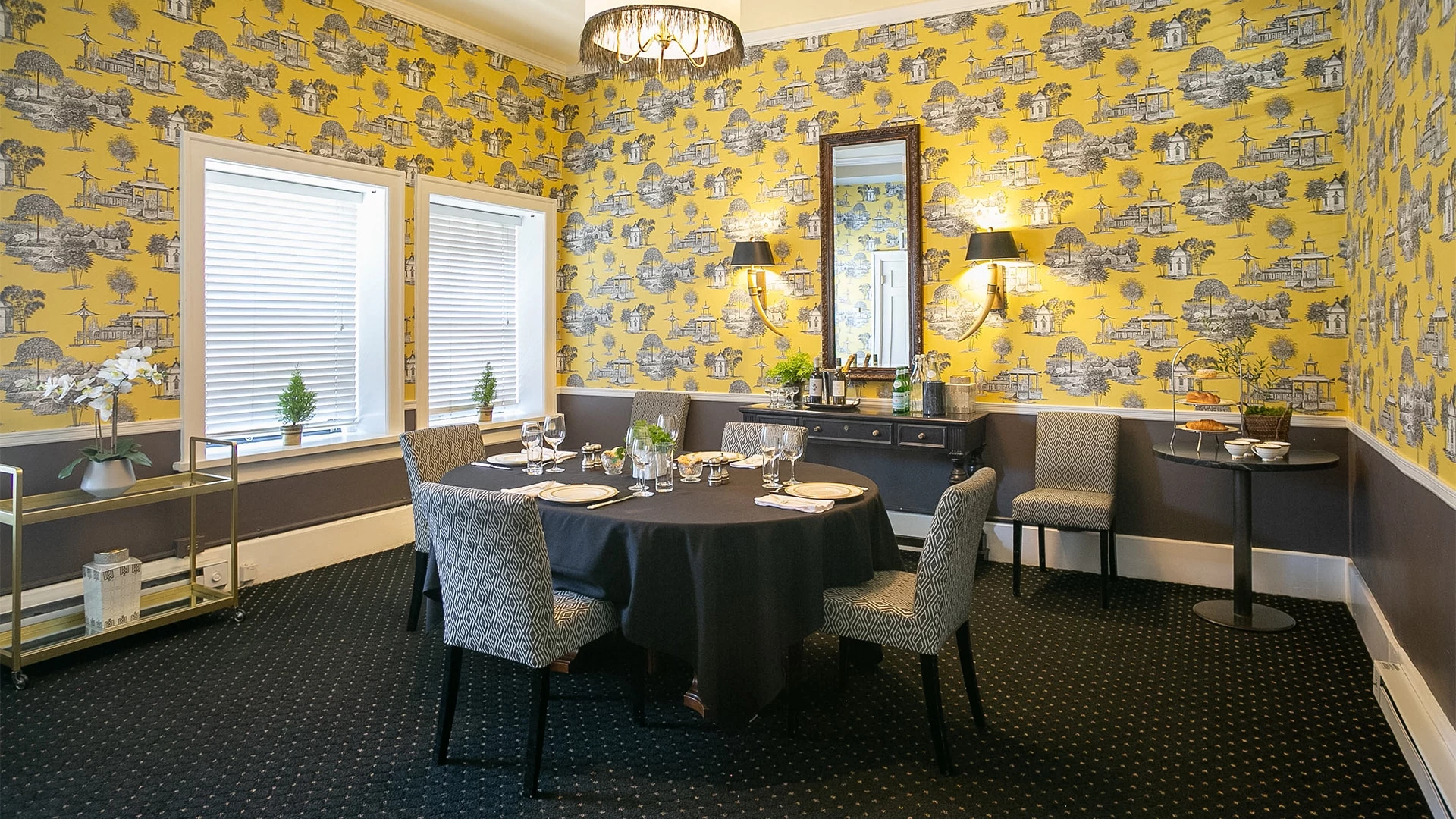 A dining room with a round table set for a meal, yellow wallpaper, and a chandelier.