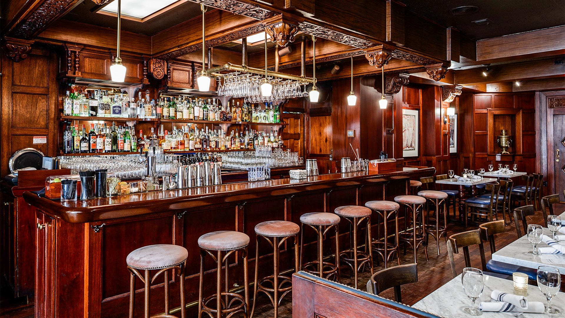 A dimly lit bar with a long wooden counter, several stools, and shelves of liquor bottles and glasses.