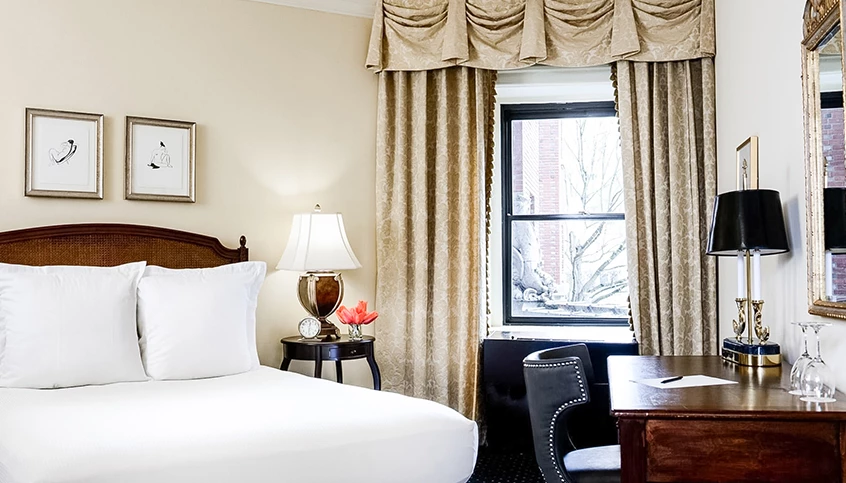 A hotel room with a bed, desk, window, and lamp.