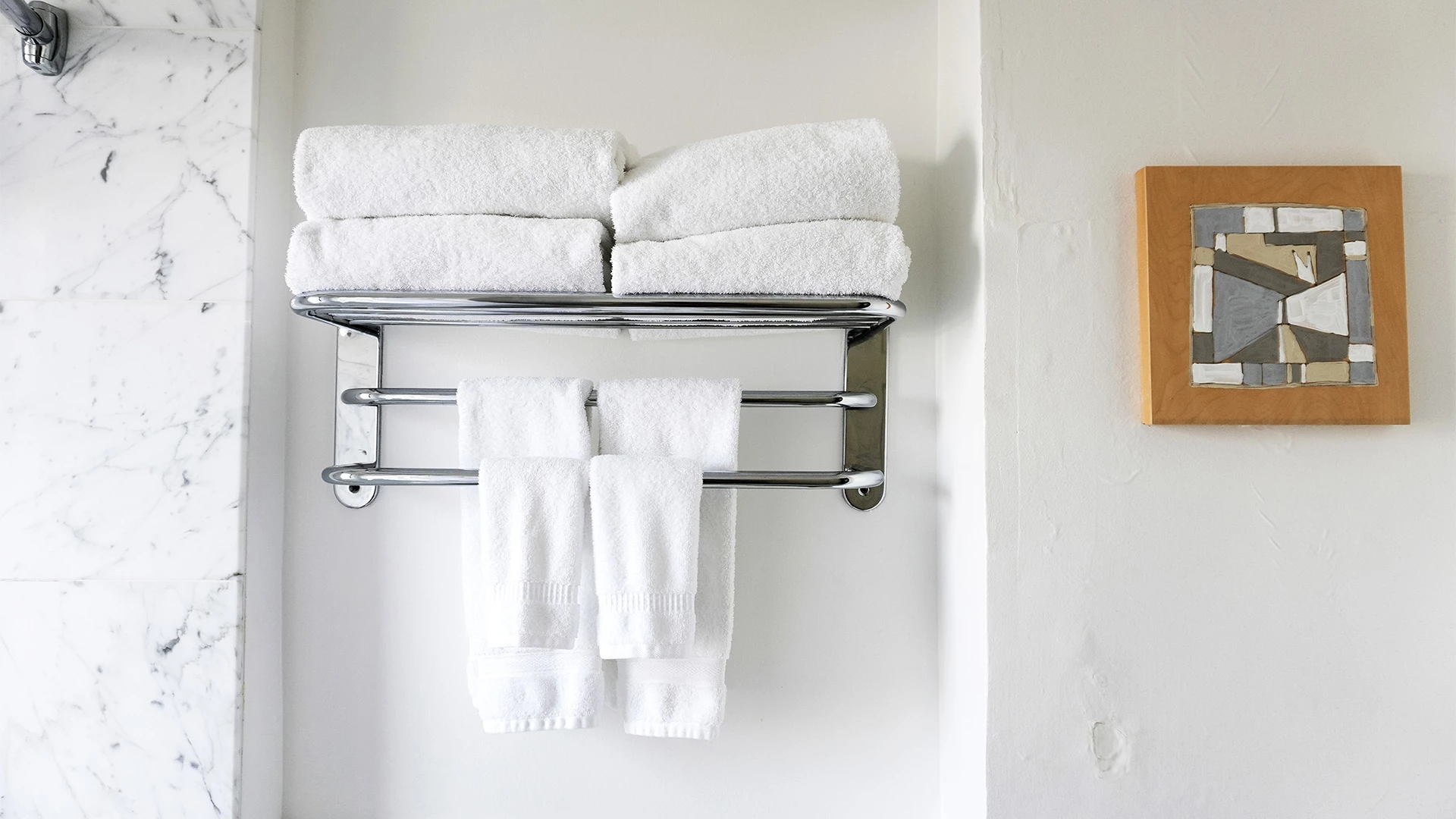 A bathroom with a towel rack holding white towels, a marble wall, and a piece of artwork.