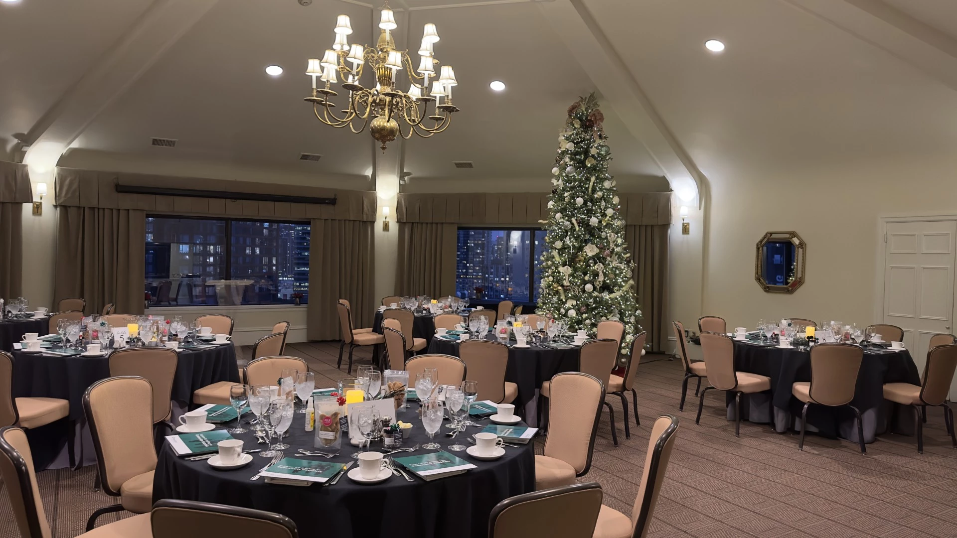 A large, elegantly set dining room with round tables, black tablecloths, and a Christmas tree.