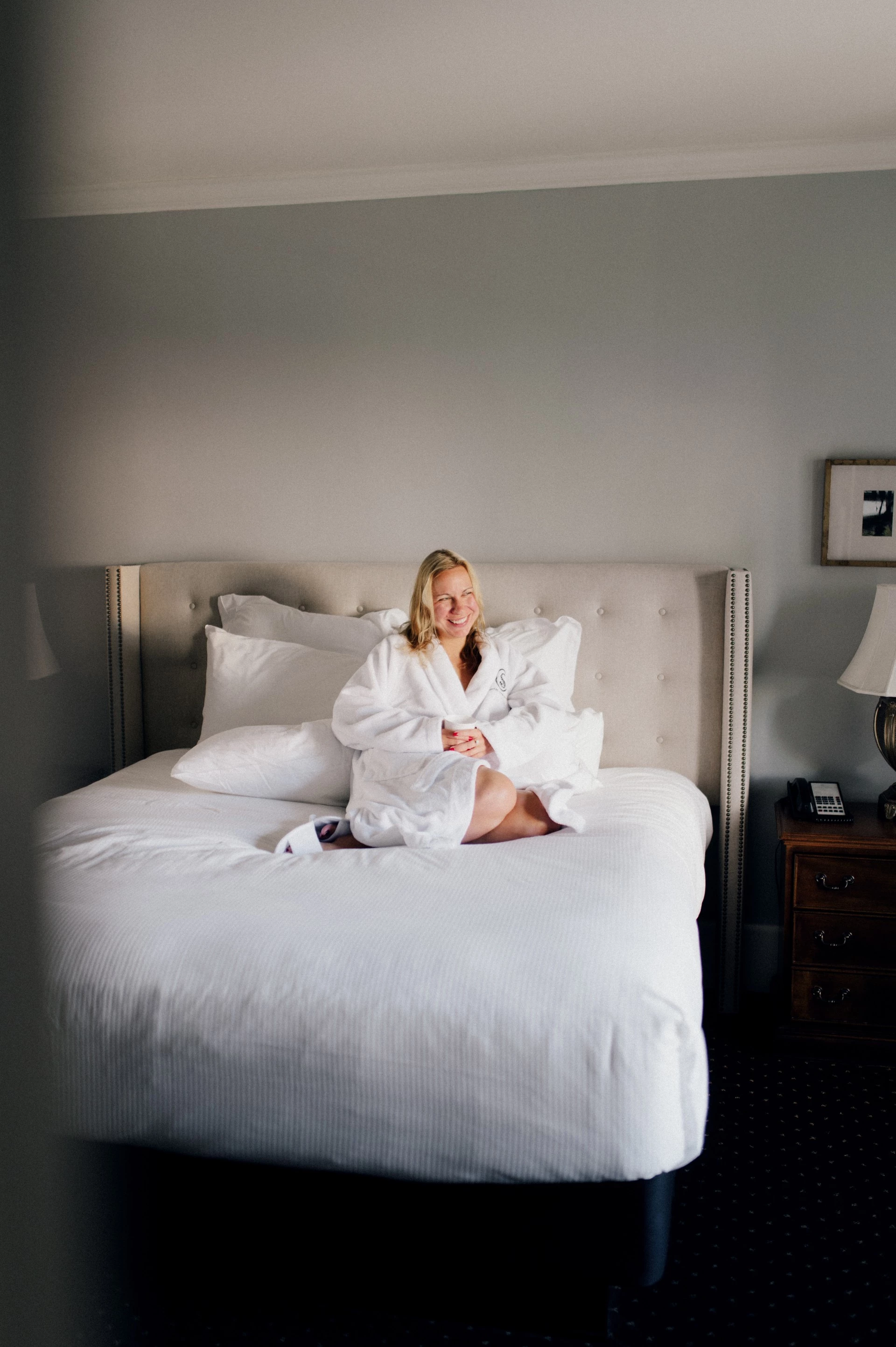 A woman in a white robe sitting on a bed in a hotel room.