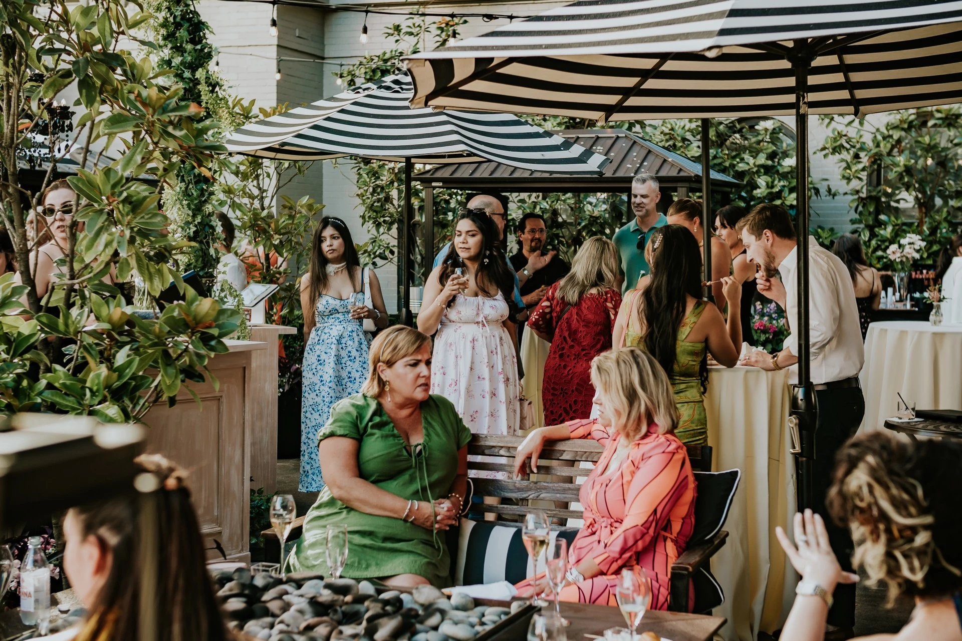 A group of people socializing in an outdoor lounge area with tables, umbrellas, and plants.