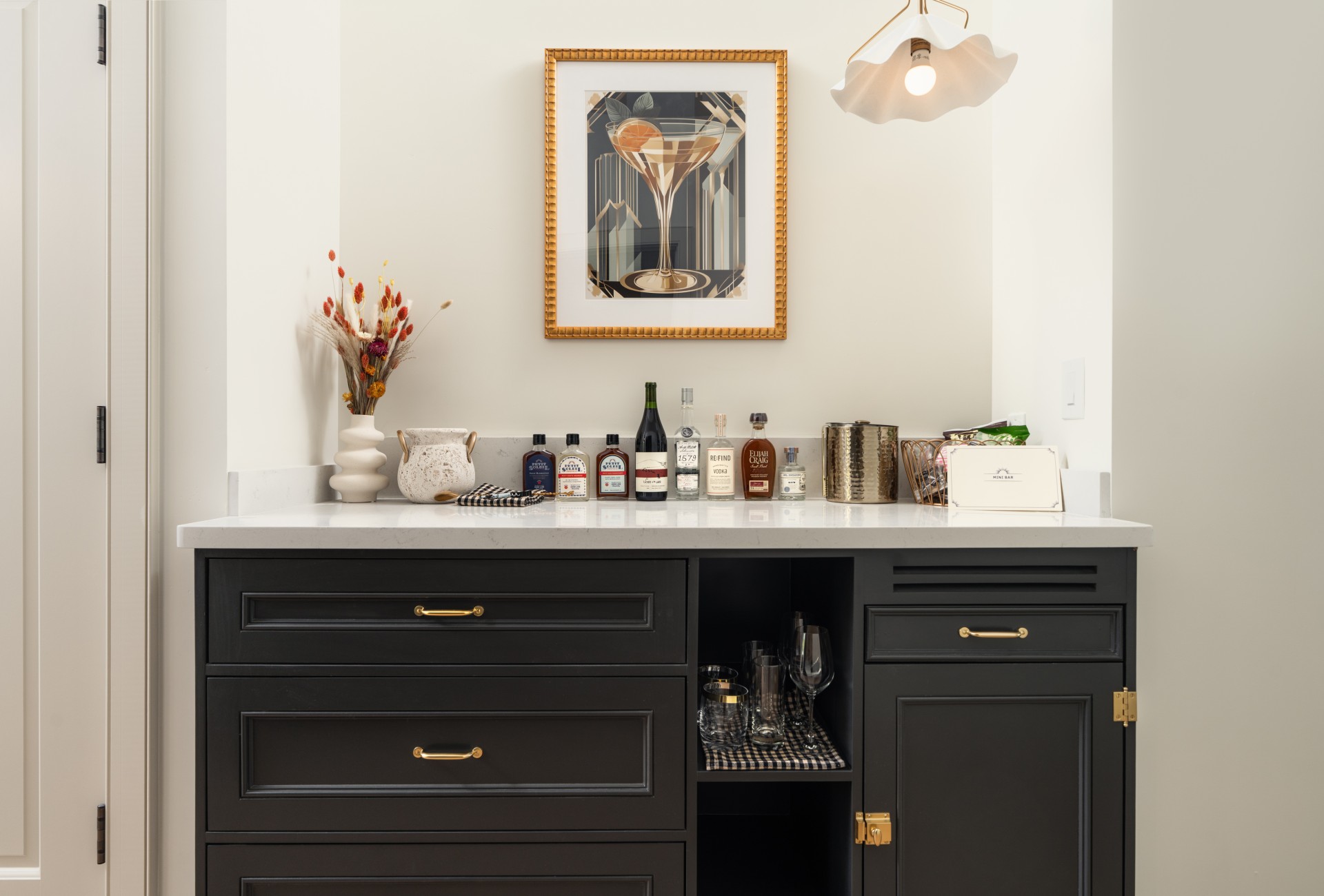 A stylish bar area with a white countertop, black cabinets with gold hardware, and a framed martini artwork above.