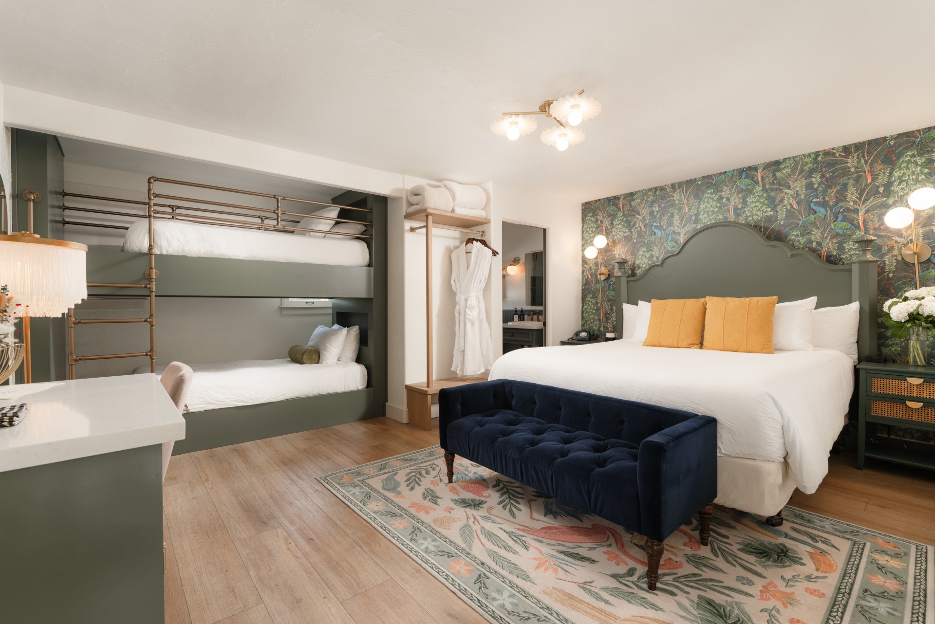 A wide view of a hotel room with a queen bed, bunk bed, velvet bench, and floral rug.