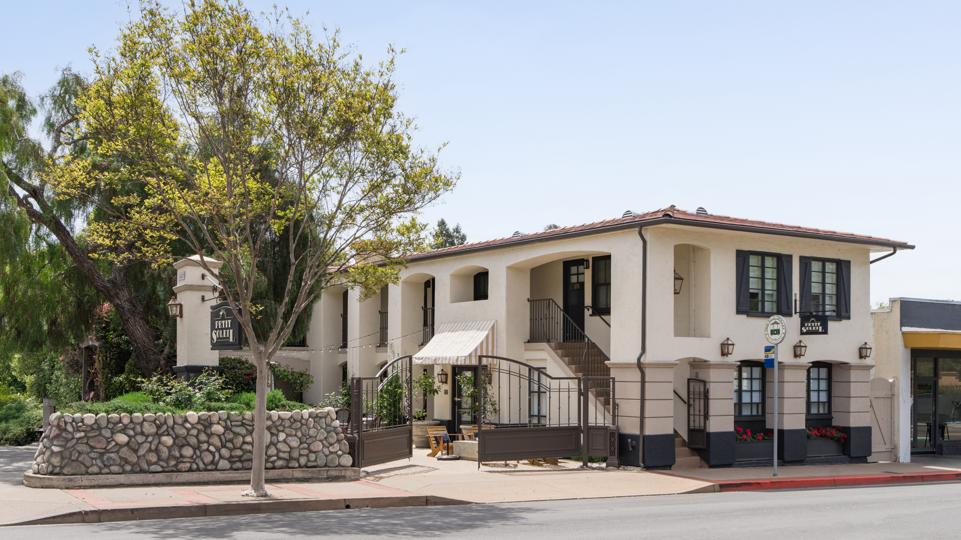 A two-story white stucco building with a red-tiled roof and black trim, featuring a stone wall and a tree in front.