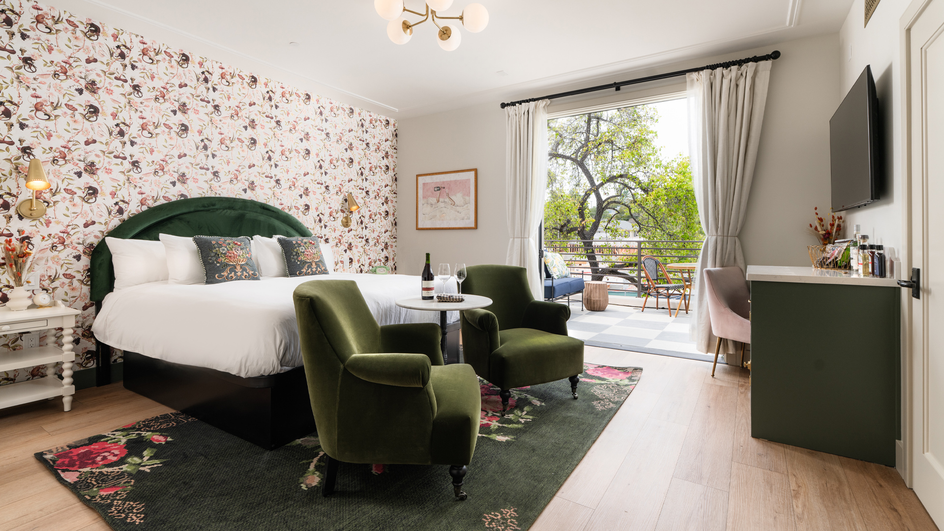 A hotel room with a green and white color scheme, floral wallpaper, and a balcony with a view of trees.