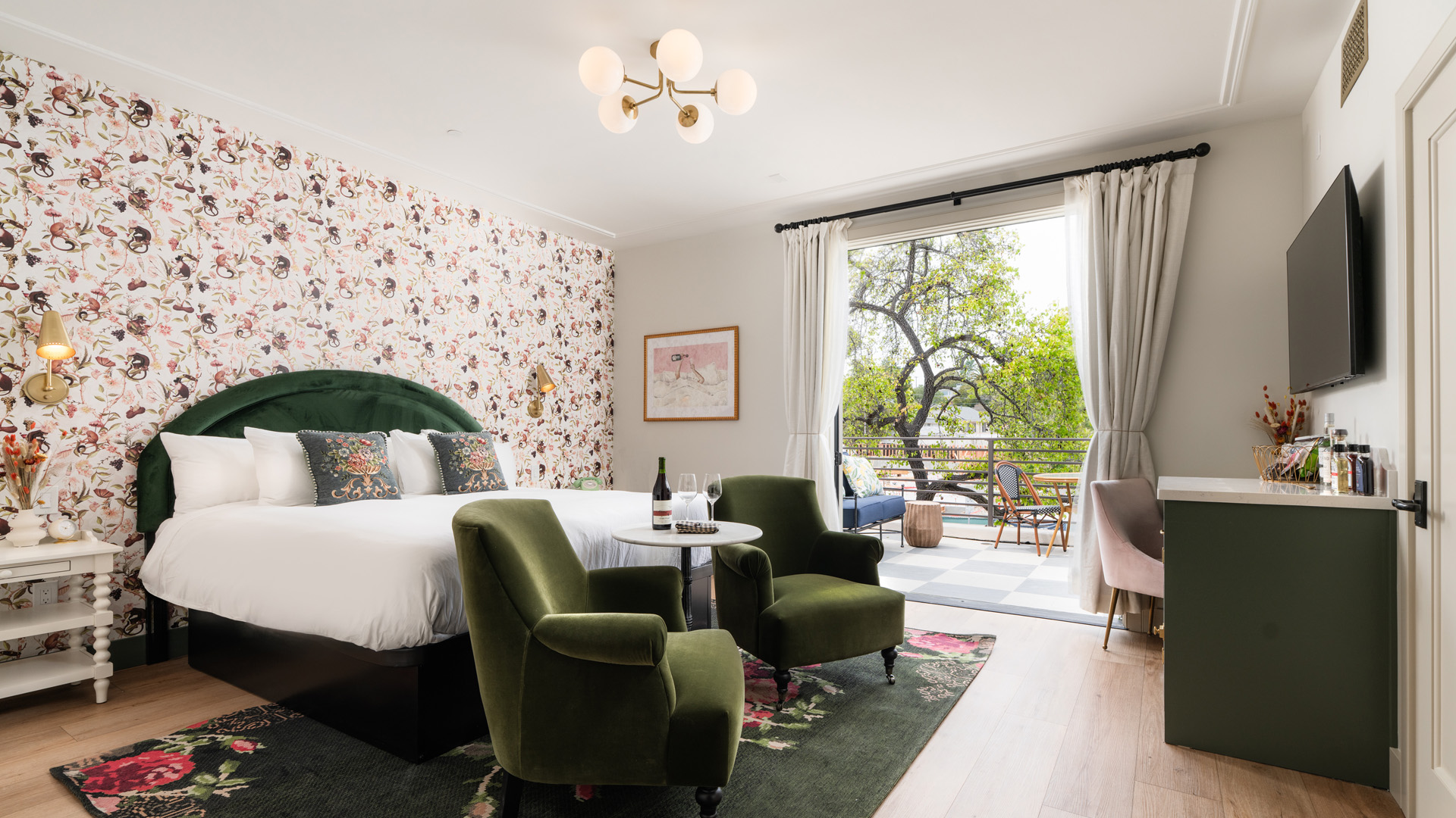 A stylish hotel room with a large bed, green armchairs, and a balcony with a view of trees.