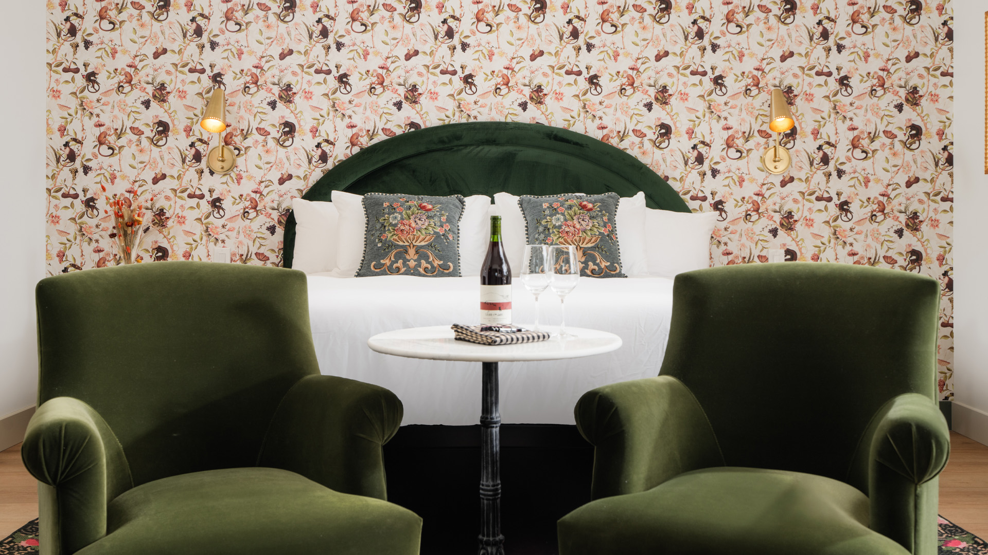 A hotel room with a bed, two green velvet chairs, a round table with a wine bottle and glasses, and floral wallpaper.