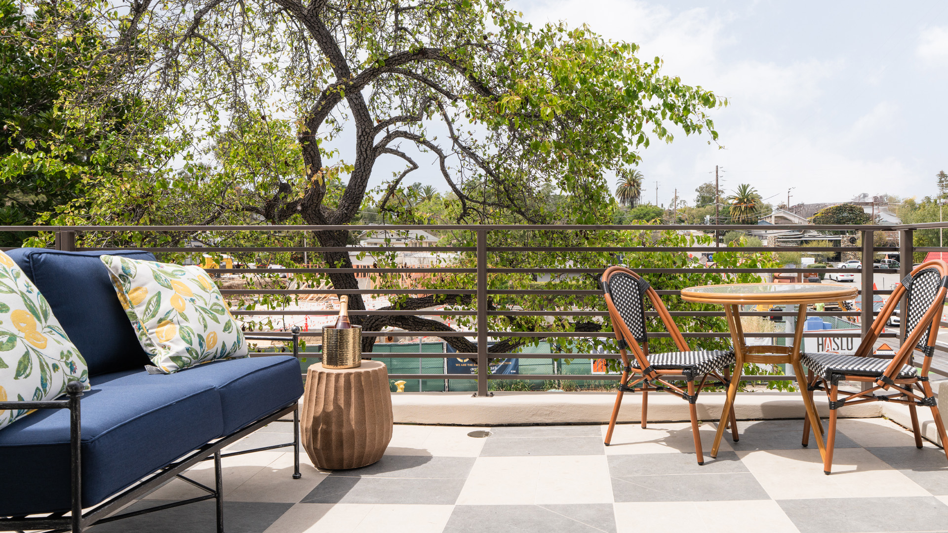 A blue sofa and wooden table with chairs on a checkered floor with a metal railing and trees in the background.