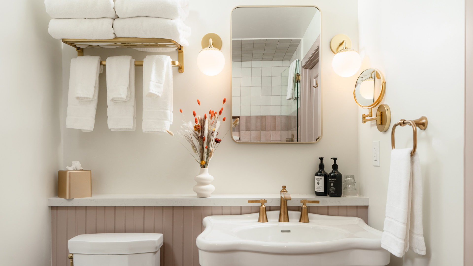A clean and stylish bathroom with a white sink, gold fixtures, and a pinkish paneling.