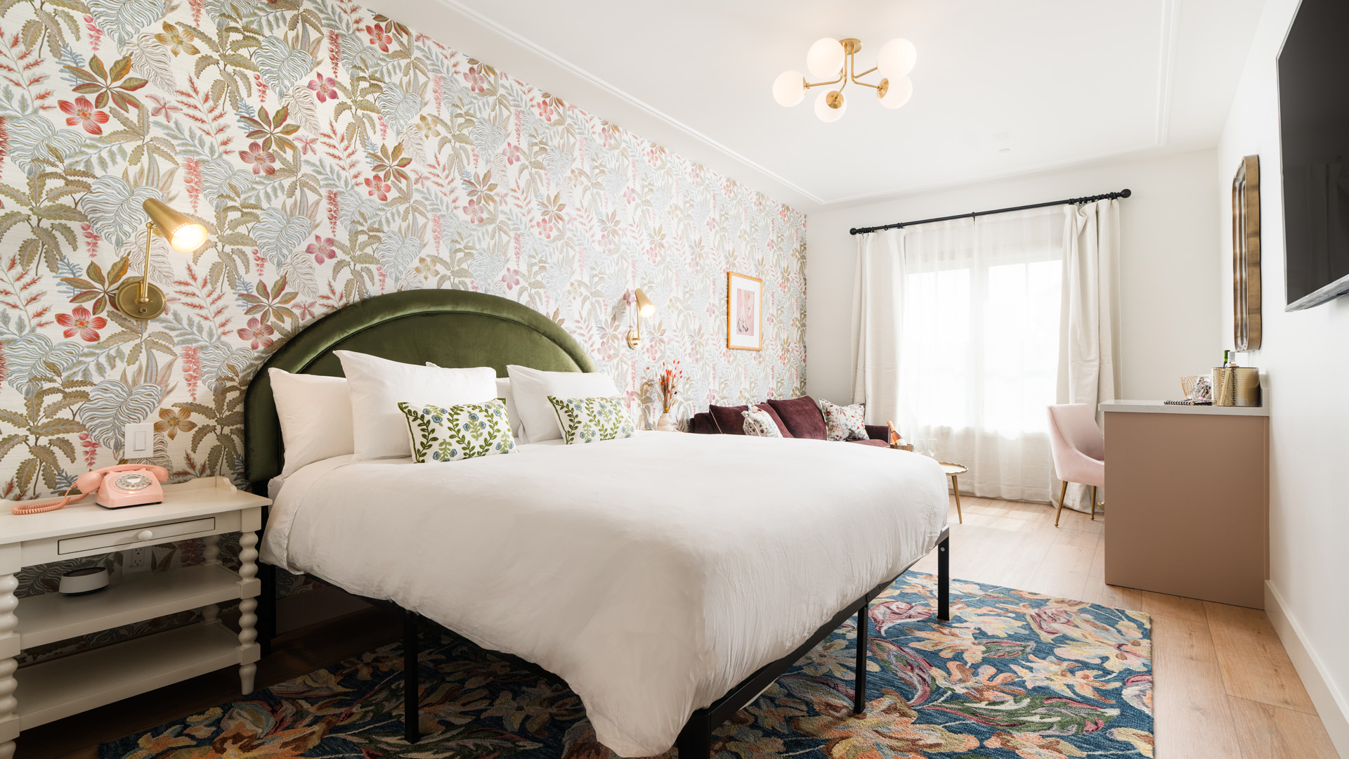 A wide-angle view of a hotel room with a floral wallpaper, a green velvet headboard, and a white bed with colorful pillows.