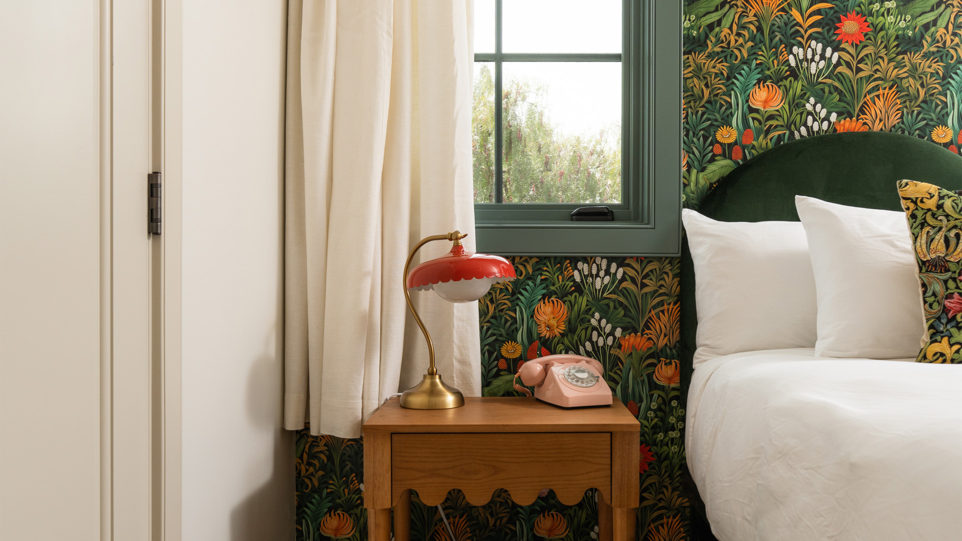 A close-up of a bed with a green headboard, white pillows, and a wooden nightstand with a red lamp and pink phone.