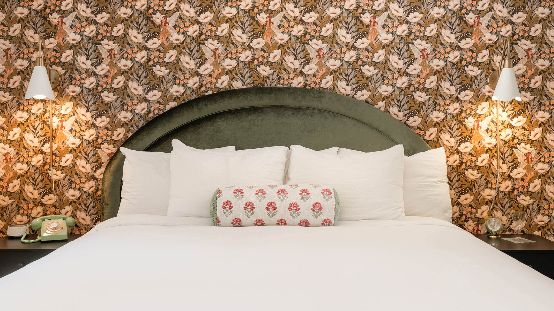 A neatly made bed with a green velvet headboard, white bedding, and floral wallpaper in a hotel room.