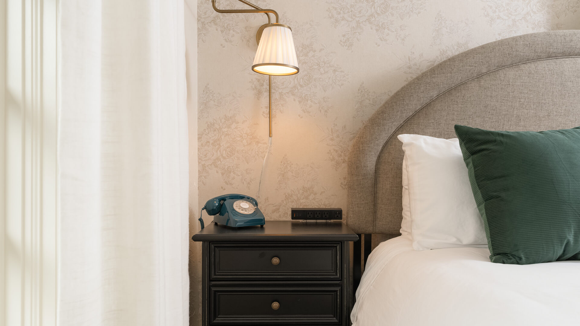 A close-up of a bed with a green pillow, white bedding, and a dark nightstand with a telephone and reading light.