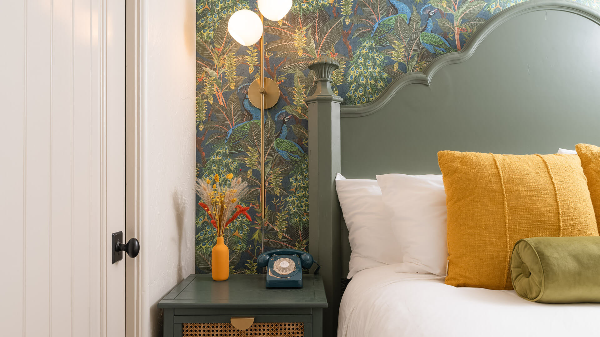 A close-up of a bed with a green headboard, yellow pillows, and a nightstand with a vase of flowers and an old phone.