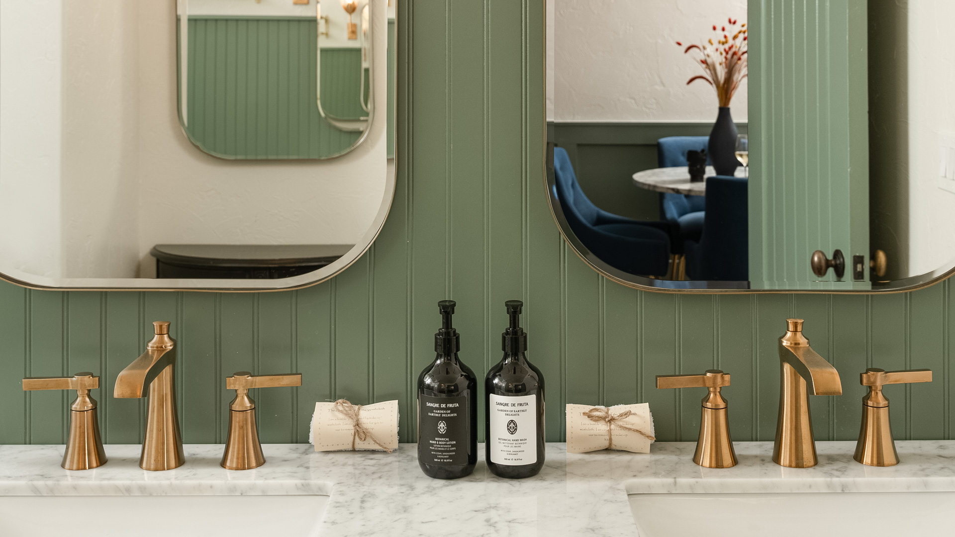 A bathroom sink area with two gold faucets, marble countertops, green paneling, and two mirrors.